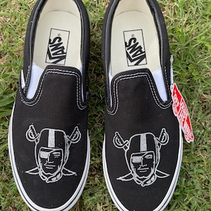 Könnte beinhalten: Paar schwarze Vans Slip-On-Schuhe mit weißer Sohle und Zierleiste. Jeder Schuh hat ein silbernes, gesticktes Raiders-Logo auf der Spitze. Das Vans-Logo ist auf der weißen Innensohle sichtbar. Ein rot-weißes Etikett ist angebracht.