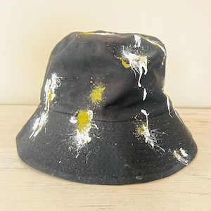 May include: Black bucket hat with white and yellow paint splatter design. The hat has a wide brim and a rounded crown. The paint splatters are randomly distributed across the hat's surface, creating a unique, artistic look.