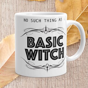May include: White ceramic mug with the text "NO SUCH THING AS A BASIC WITCH" in black, with decorative flourishes. The mug is on a wooden surface with yellow and brown leaves.