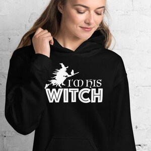 以下が含まれることがあります： 黒色のフード付きスウェットシャツ。白い魔女がほうきに乗っているグラフィックと「I'M HIS WITCH」の文字がプリントされています。スウェットシャツにはフロントポケットとドローストリングフードが付いています。デザインは前面の中央に配置されています。