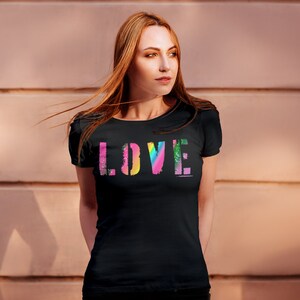 Puede incluir: Camiseta negra con la palabra "LOVE" en un dise&ntilde;o colorido tipo arco&iacute;ris. La palabra est&aacute; impresa en una fuente audaz y en bloque, con cada letra que presenta un degradado de color diferente. La camiseta tiene mangas cortas y cuello redondo.