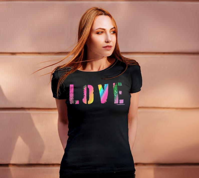 Puede incluir: Camiseta negra con la palabra "LOVE" en un dise&ntilde;o colorido tipo arco&iacute;ris. La palabra est&aacute; impresa en una fuente audaz y en bloque, con cada letra que presenta un degradado de color diferente. La camiseta tiene mangas cortas y cuello redondo.