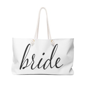 May include: White tote bag with the word "bride" in black script. The bag has white rope handles and gold-colored accents. A large, casual bag for carrying items.