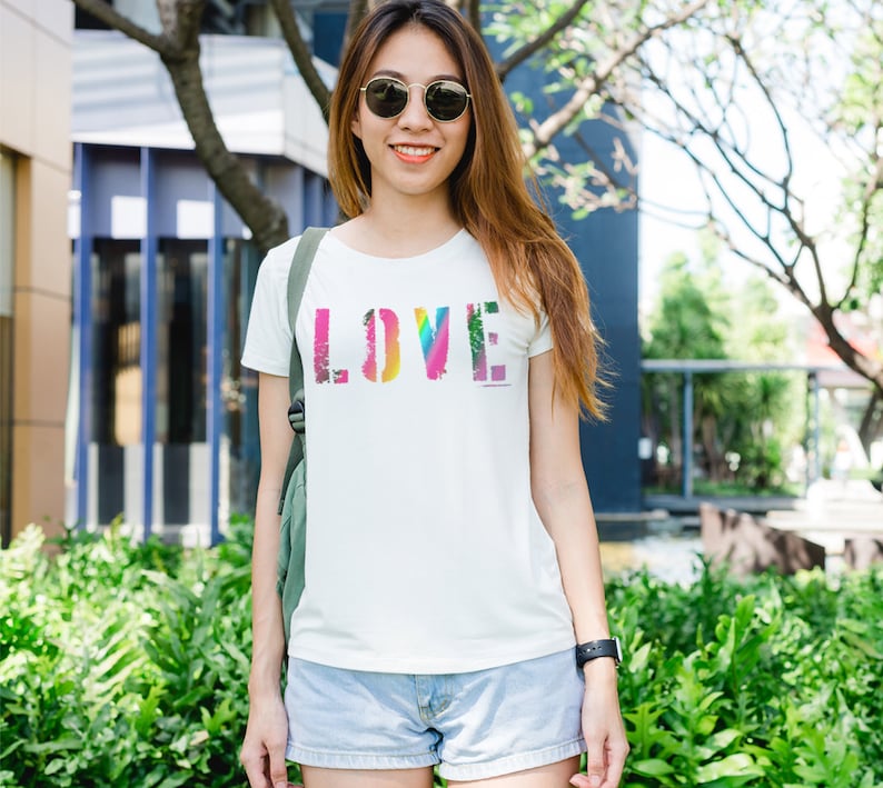 Puede incluir: Una camiseta blanca con la palabra "LOVE" impresa en letras multicolores y desgastadas. La persona lleva gafas de sol y est&aacute; al aire libre.