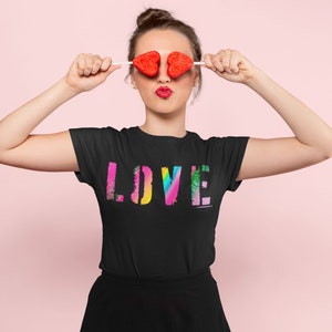 Puede incluir: Camiseta negra con la palabra "LOVE" en un dise&ntilde;o colorido y texturizado. La persona de la imagen sostiene piruletas en forma de coraz&oacute;n sobre sus ojos. El fondo es de color rosa liso.