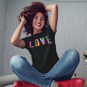 Puede incluir: Camiseta negra con la palabra "LOVE" en un dise&ntilde;o colorido, similar a un arco&iacute;ris. La camiseta la lleva una persona sentada con los brazos levantados. La persona lleva vaqueros azules y zapatos rojos.