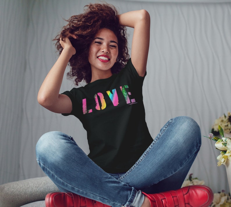 Puede incluir: Camiseta negra con la palabra "LOVE" en un dise&ntilde;o colorido, similar a un arco&iacute;ris. La camiseta la lleva una persona sentada con los brazos levantados. La persona lleva vaqueros azules y zapatos rojos.