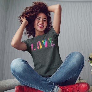 Puede incluir: Una camiseta gris oscuro con la palabra "LOVE" en un dise&ntilde;o colorido, similar a un arco&iacute;ris. La persona lleva vaqueros azules y zapatos rojos. La persona est&aacute; sentada con los brazos levantados.