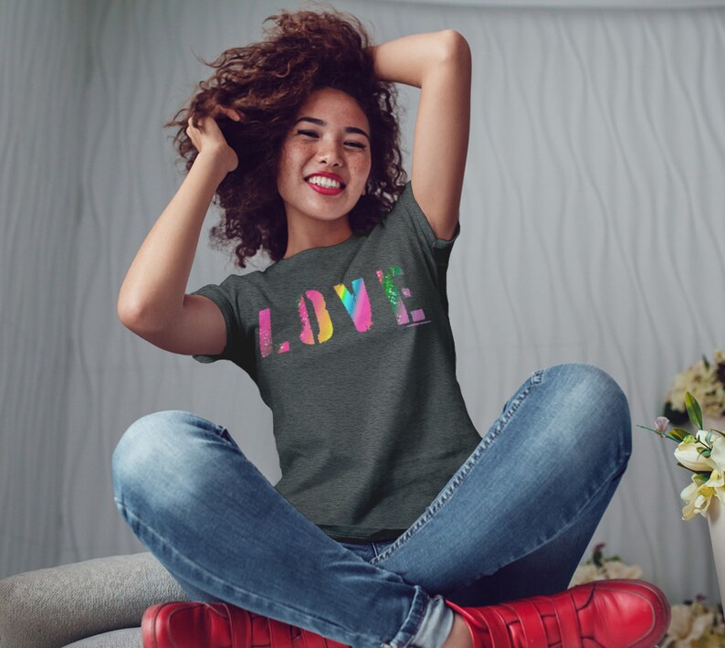 Puede incluir: Una camiseta gris oscuro con la palabra "LOVE" en un dise&ntilde;o colorido, similar a un arco&iacute;ris. La persona lleva vaqueros azules y zapatos rojos. La persona est&aacute; sentada con los brazos levantados.