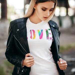 Puede incluir: Camiseta blanca con la palabra "LOVE" en fuente desgastada de color arco&iacute;ris. La camiseta se lleva debajo de una chaqueta de cuero negra. La imagen est&aacute; tomada al aire libre con un fondo borroso.