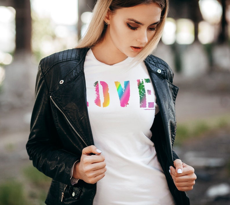 Puede incluir: Camiseta blanca con la palabra "LOVE" en fuente desgastada de color arco&iacute;ris. La camiseta se lleva debajo de una chaqueta de cuero negra. La imagen est&aacute; tomada al aire libre con un fondo borroso.