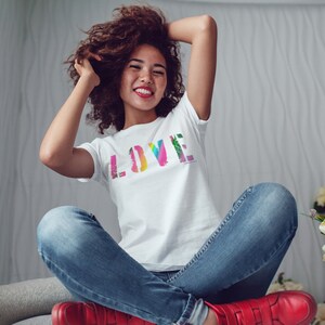 Puede incluir: Una camiseta blanca con la palabra "LOVE" en un dise&ntilde;o colorido, similar a un arco&iacute;ris. La camiseta la lleva una persona con vaqueros azules y zapatos rojos. La persona sonr&iacute;e y tiene los brazos levantados.