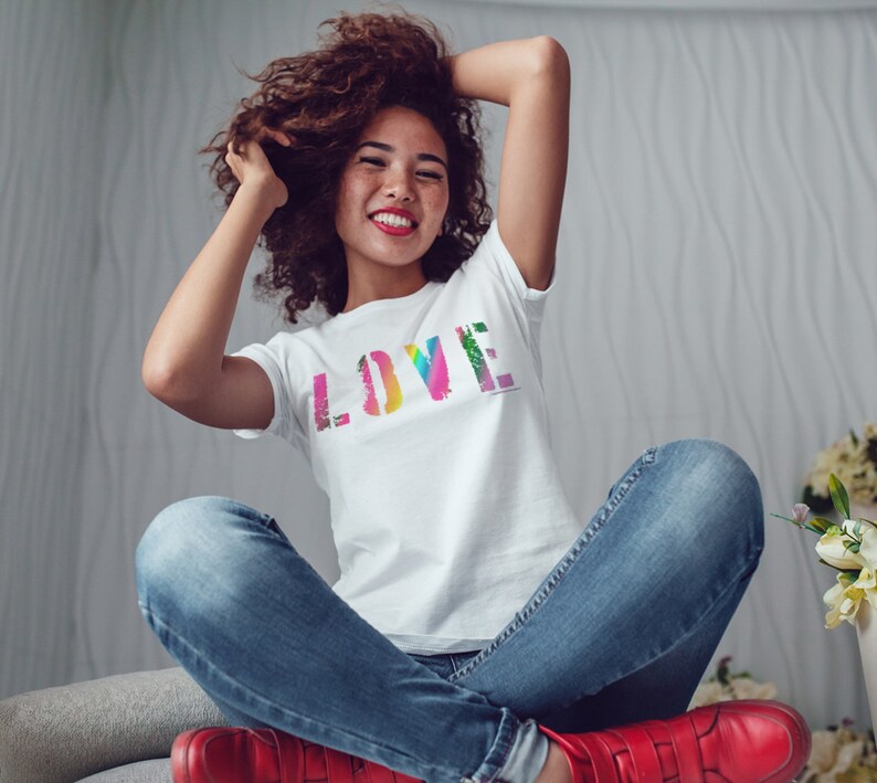 Puede incluir: Una camiseta blanca con la palabra "LOVE" en un dise&ntilde;o colorido, similar a un arco&iacute;ris. La camiseta la lleva una persona con vaqueros azules y zapatos rojos. La persona sonr&iacute;e y tiene los brazos levantados.