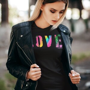 Puede incluir: Camiseta negra con la palabra "LOVE" en un dise&ntilde;o colorido y desgastado de arco&iacute;ris. La persona lleva una chaqueta de cuero negra.