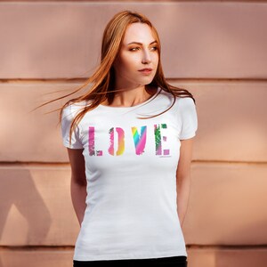 Puede incluir: Camiseta blanca con la palabra "LOVE" en una fuente colorida y desgastada en el pecho. Las letras son una mezcla de tonos arco&iacute;ris, incluyendo rosa, amarillo, verde y azul. La camiseta tiene mangas cortas y cuello redondo.