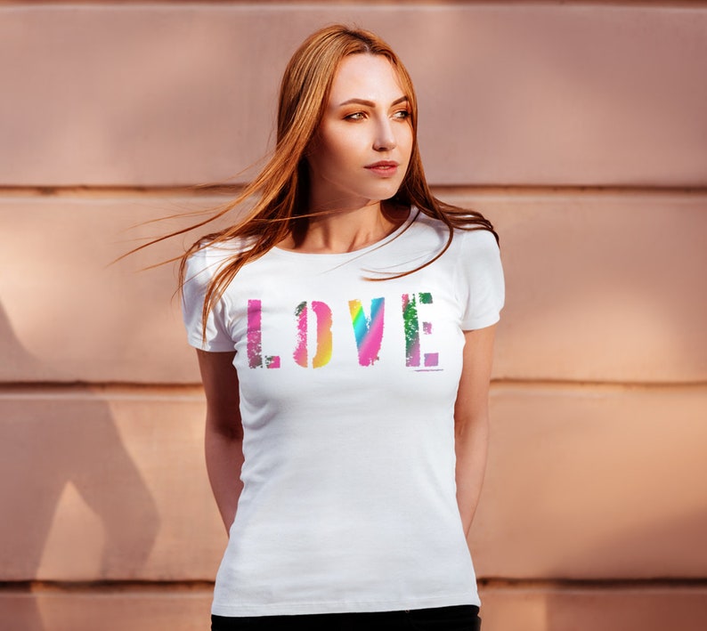 Puede incluir: Camiseta blanca con la palabra "LOVE" en una fuente colorida y desgastada en el pecho. Las letras son una mezcla de tonos arco&iacute;ris, incluyendo rosa, amarillo, verde y azul. La camiseta tiene mangas cortas y cuello redondo.