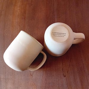 May include: Two white ceramic mugs with rounded edges and handles. The mug on the right has a black label that reads "by ANTHROPOLOGIE  Fabriqué au Portugal/Made in Portugal  Dishwasher and Microwave safe."