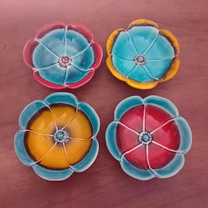 Può includere: Quattro ciotole in ceramica a forma di fiore nei toni del blu, rosso e giallo. Ogni ciotola ha un motivo floreale centrale e un bordo smerlato. Le ciotole sono disposte su una superficie marrone.