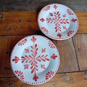 Puede incluir: Dos platos de cerámica blanca con un diseño floral rojo. Los platos tienen un patrón central en forma de copo de nieve y un borde punteado. Están sobre una superficie de madera.