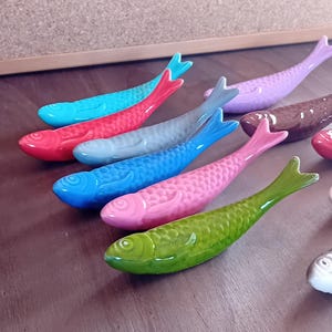 Op de afbeelding: Een set kleurrijke keramische visfiguren in verschillende tinten blauw, groen, roze, rood, paars en bruin. De vissen zijn in een rij op een houten oppervlak gerangschikt.