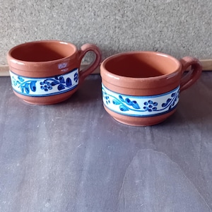 Può includere: Due piccole tazze in ceramica color terracotta con manici. Ogni tazza presenta una fascia bianca con un motivo floreale blu sul bordo. Le tazze sono esposte su una superficie di legno.