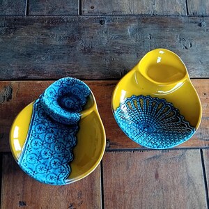 May include: Two ceramic serving dishes. Each dish is yellow with a blue decorative design. The dishes have a unique shape with a small, raised section on one side. The blue design is a lace-like pattern.