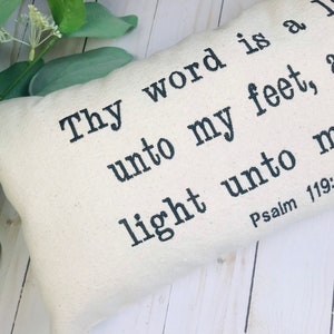 Puede incluir: Coj&iacute;n rectangular crema con texto negro. El texto dice: "L&aacute;mpara es a mis pies tu palabra, y luz para mi camino. Salmo 119:105". El coj&iacute;n est&aacute; sobre una superficie de madera blanca con follaje verde.