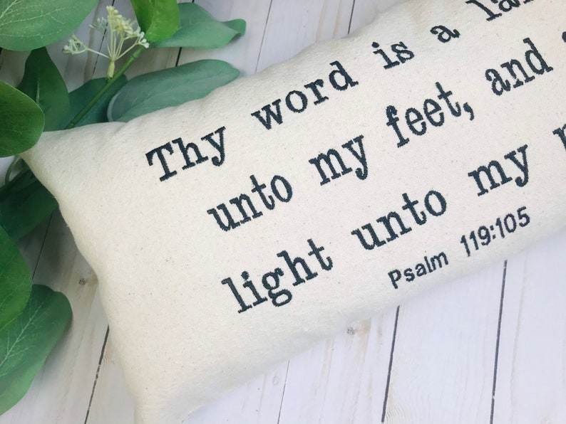 Puede incluir: Coj&iacute;n rectangular crema con texto negro. El texto dice: "L&aacute;mpara es a mis pies tu palabra, y luz para mi camino. Salmo 119:105". El coj&iacute;n est&aacute; sobre una superficie de madera blanca con follaje verde.