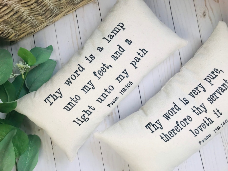 Puede incluir: Dos almohadas rectangulares decorativas de color blanquecino con texto negro. Una almohada dice: "L&aacute;mpara es a mis pies tu palabra, y lumbrera a mi camino Salmo 119:105". La otra dice: "Mucho es tu palabra amada de tu siervo Salmo 119:140."