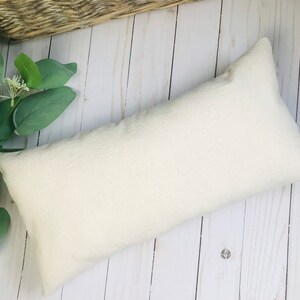 Puede incluir: Una almohada rectangular blanquecina con una superficie texturizada. La almohada est&aacute; colocada sobre una superficie de madera clara, con follaje verde y una cesta tejida en el fondo. La forma de la almohada es alargada.