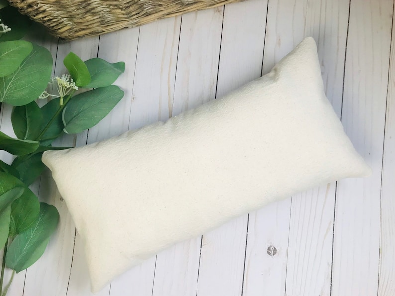 Puede incluir: Una almohada rectangular blanquecina con una superficie texturizada. La almohada est&aacute; colocada sobre una superficie de madera clara, con follaje verde y una cesta tejida en el fondo. La forma de la almohada es alargada.