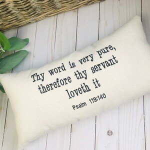 Puede incluir: Coj&iacute;n rectangular color crema con el texto "Thy word is very pure, therefore thy servant loveth it Psalm 119:140" impreso en negro. El coj&iacute;n est&aacute; sobre una superficie de madera clara, con vegetaci&oacute;n y una cesta de mimbre en el fondo.