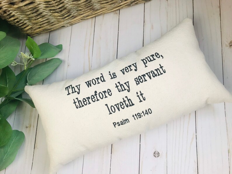 Puede incluir: Coj&iacute;n rectangular color crema con el texto "Thy word is very pure, therefore thy servant loveth it Psalm 119:140" impreso en negro. El coj&iacute;n est&aacute; sobre una superficie de madera clara, con vegetaci&oacute;n y una cesta de mimbre en el fondo.