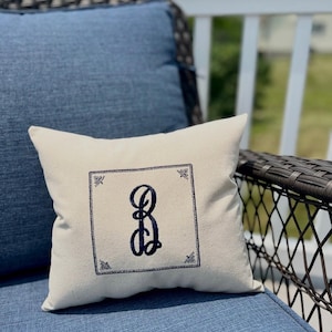 May include: A square, off-white throw pillow with a navy blue embroidered letter "G" inside a decorative square border. The pillow is resting on a blue cushion, with a wicker chair and white railing in the background.