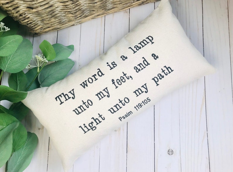 Puede incluir: Coj&iacute;n rectangular color crema con texto negro: "Tu palabra es una l&aacute;mpara a mis pies, y una luz en mi camino Salmo 119:105." El coj&iacute;n est&aacute; sobre una superficie de madera blanca con vegetaci&oacute;n y una cesta tejida en el fondo.