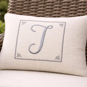 May include: Cream-colored decorative pillow with a gray embroidered letter "J" inside a square frame. The pillow is on a wicker chair, suggesting outdoor or patio use. The design includes corner embellishments.