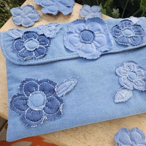 Puede incluir: Un bolso de mano tipo sobre hecho de tela vaquera azul claro con múltiples aplicaciones de flores de tela en varios tonos de azul. Las flores están cosidas en la solapa frontal y el cuerpo del bolso, con bordes deshilachados y costuras blancas.