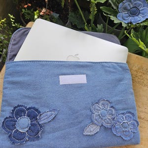 Puede incluir: Una funda para portátil de tela vaquera azul claro con un cierre de velcro rectangular blanco y tres aplicaciones de flores de tela en varios tonos de azul. La funda está parcialmente abierta, dejando ver un portátil plateado.