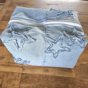 May include: Light blue denim pouch with white zipper and star cutouts. The pouch is made of denim and features several star-shaped cutouts. The zipper is white, and the pouch is sitting on a wooden surface.