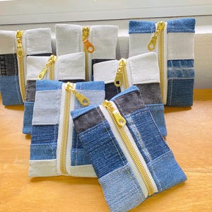 Puede incluir: Una colección de bolsas de patchwork de mezclilla con cremalleras doradas. Las bolsas son rectangulares, con una variedad de tonos de mezclilla azul y secciones de tela blanca. Las cremalleras recorren la parte superior, sobre una superficie de madera.