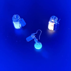 May include: Three small glass jars with cork tops and silver tags that read "Drink Me" hanging from silver earring hooks. The jars are filled with a white substance that glows under a black light.