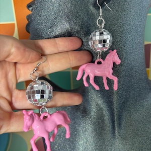 May include: A pair of drop earrings featuring a silver-toned metal hook, a spherical disco ball charm covered in small reflective mirror tiles, and a solid light pink plastic horse figurine suspended from the bottom of each disco ball.