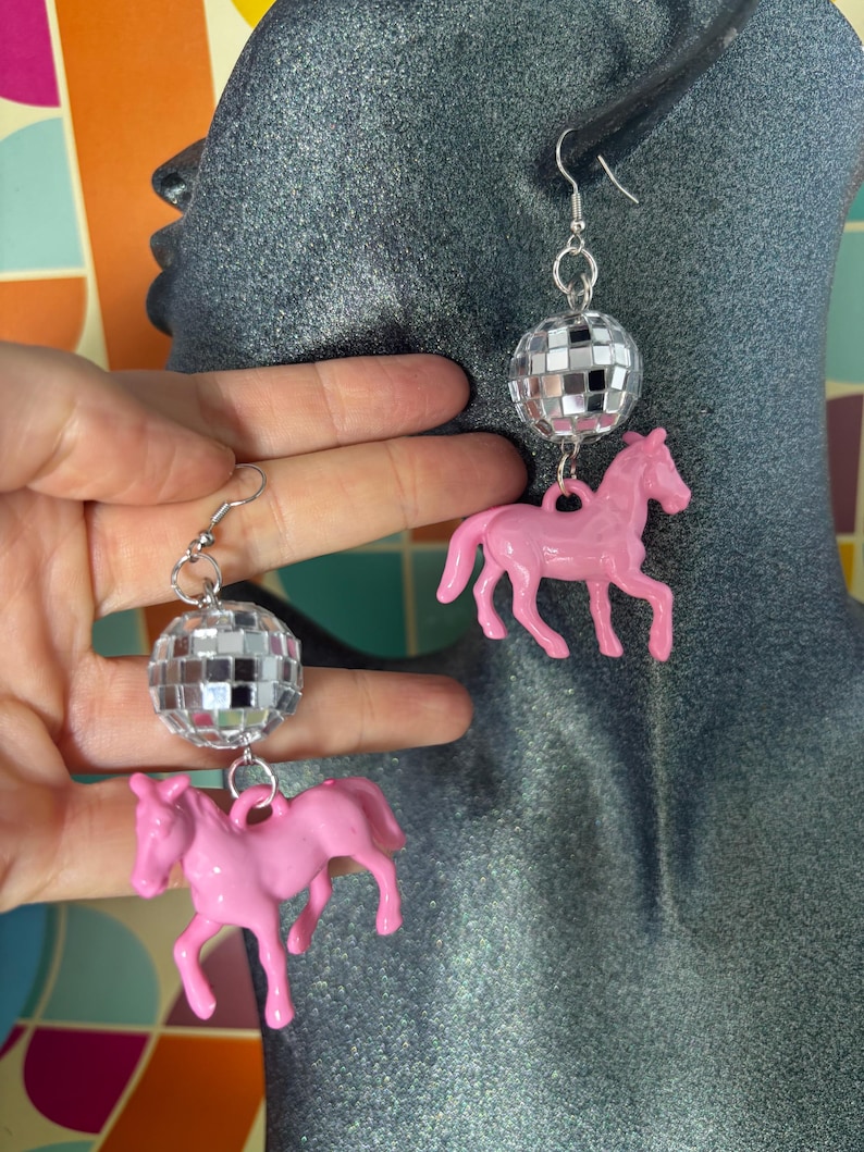 May include: A pair of drop earrings featuring a silver-toned metal hook, a spherical disco ball charm covered in small reflective mirror tiles, and a solid light pink plastic horse figurine suspended from the bottom of each disco ball.