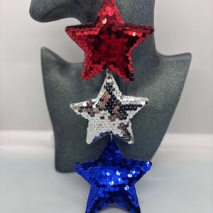 May include: Dangling earrings featuring three sequined stars in red, silver, and blue. The stars are layered, with the red star at the top, followed by a silver star, and a blue star at the bottom. The earrings are hanging from a silver hook.