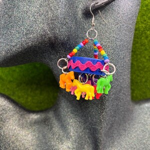 May include: A dangle earring featuring a blue plastic bottle cap wrapped in pink rickrack trim. The earring is suspended by a triangle of multicolored beads and features several small, colorful plastic horse charms hanging from silver metal rings.