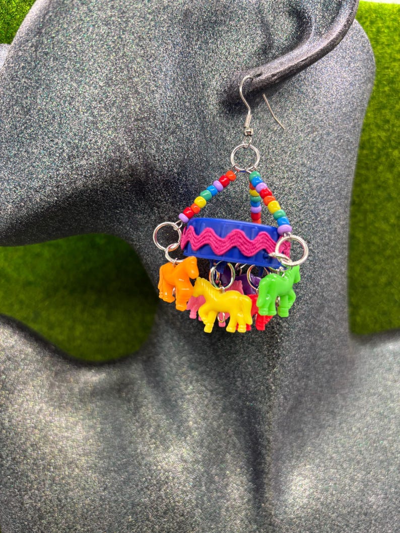 May include: A dangle earring featuring a blue plastic bottle cap wrapped in pink rickrack trim. The earring is suspended by a triangle of multicolored beads and features several small, colorful plastic horse charms hanging from silver metal rings.