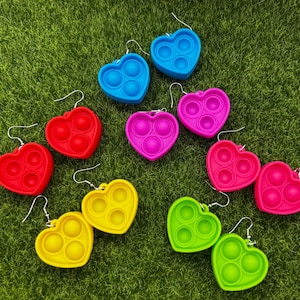 May include: A collection of heart-shaped earrings in various colors, including red, blue, pink, yellow, and green. Each earring features a bubble-popping design with three circular indentations. The earrings are displayed on a green surface.