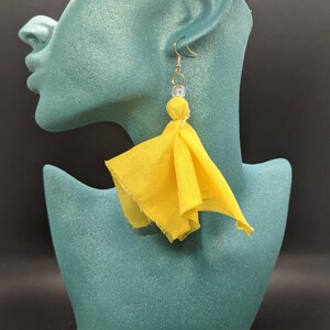 Puede incluir: Pendientes de borla de tela amarilla con un gancho dorado y una cuenta transparente. Los pendientes cuelgan de la oreja de un maniqu&iacute;.