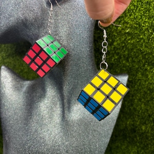 Könnte beinhalten: Ein Paar ausgefallene Ohrringe mit Miniatur-Rubik's-Würfeln. Ein Ohrring zeigt rote und grüne Quadrate, der andere gelbe und blaue. Sie hängen an silberfarbenen Kettengliedern und Haken.