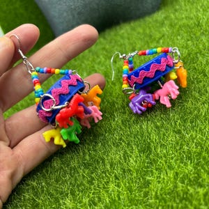May include: A pair of dangle earrings featuring a blue plastic bottle cap base decorated with pink rickrack trim. The earrings are suspended by multi-colored rainbow beads and feature small, colorful plastic horse charms in shades of red, yellow, green, orange, and purple hanging from the bottom.